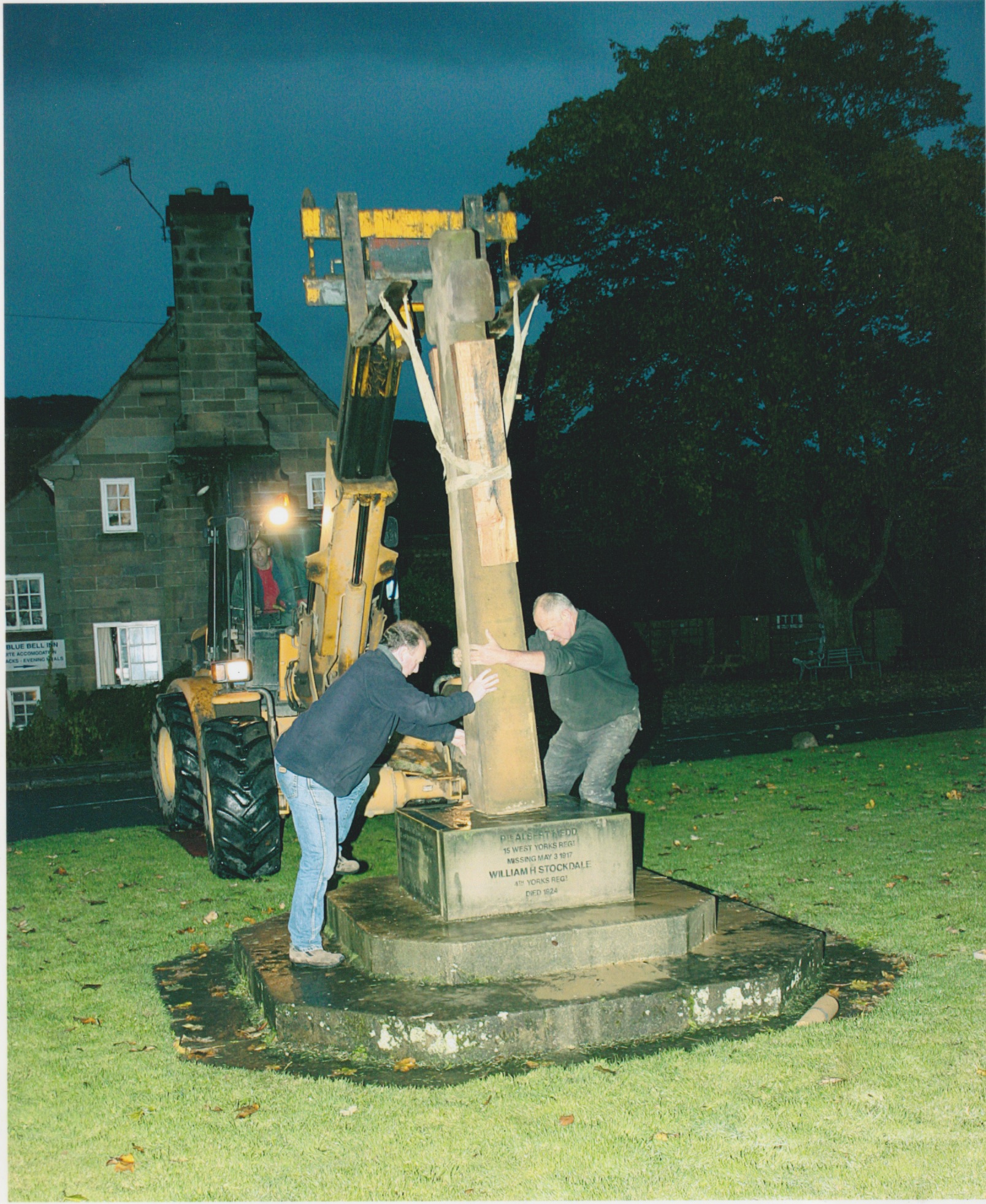 two men holding the war memorial, as a man in a tractor raises it to place to be fixed.